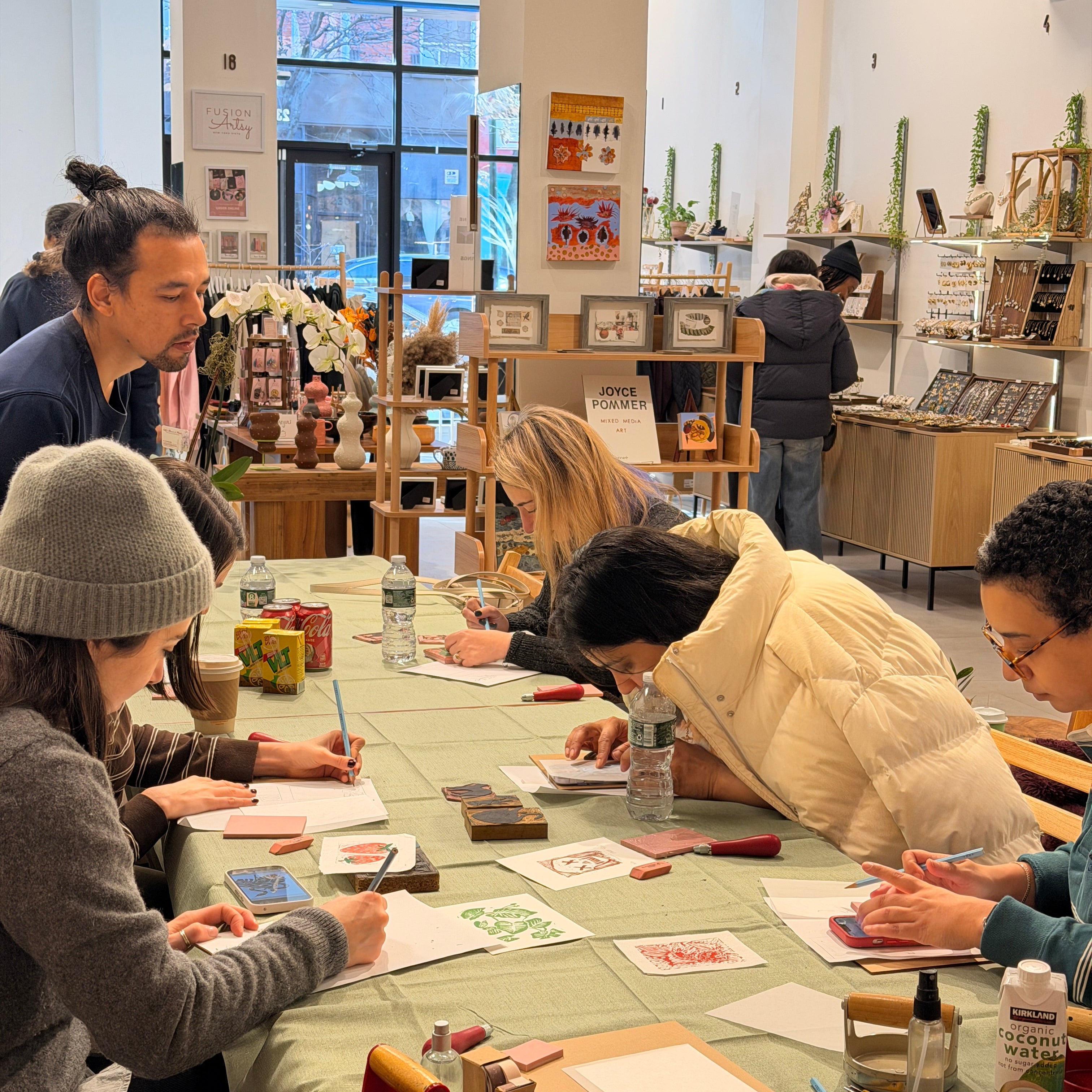 A group of people sitting around a table in a bright shop, painting and stamping on paper during what looks like a workshop or class. The table is covered with art supplies, including paintbrushes, canvases, and stamps.