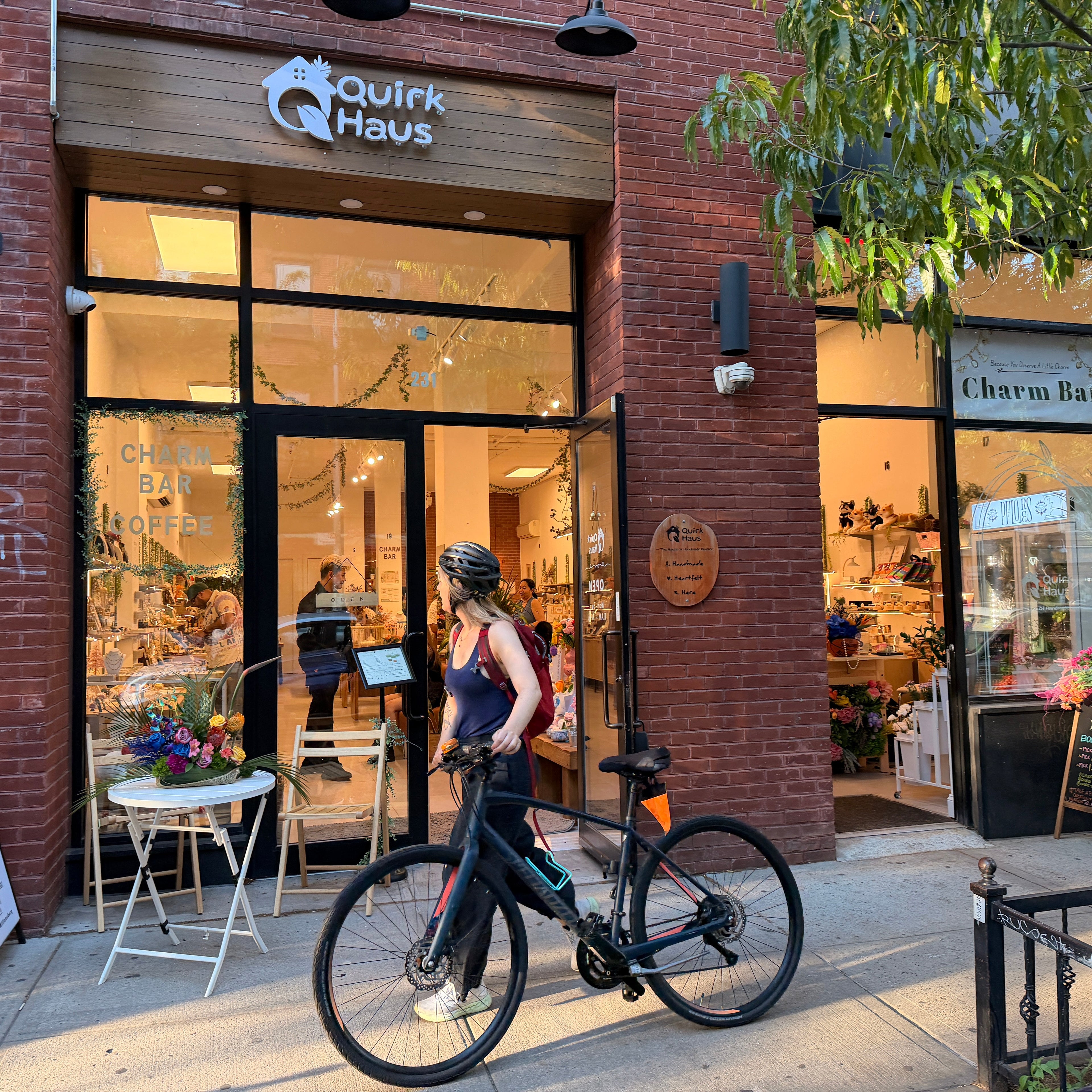 A woman wearing a helmet stands on a city sidewalk next to a black bicycle, with the Quik Haus storefront behind her. The brick shop front has large glass windows, interior lighting, outdoor seating, and a second storefront sign reading Charm Bar.