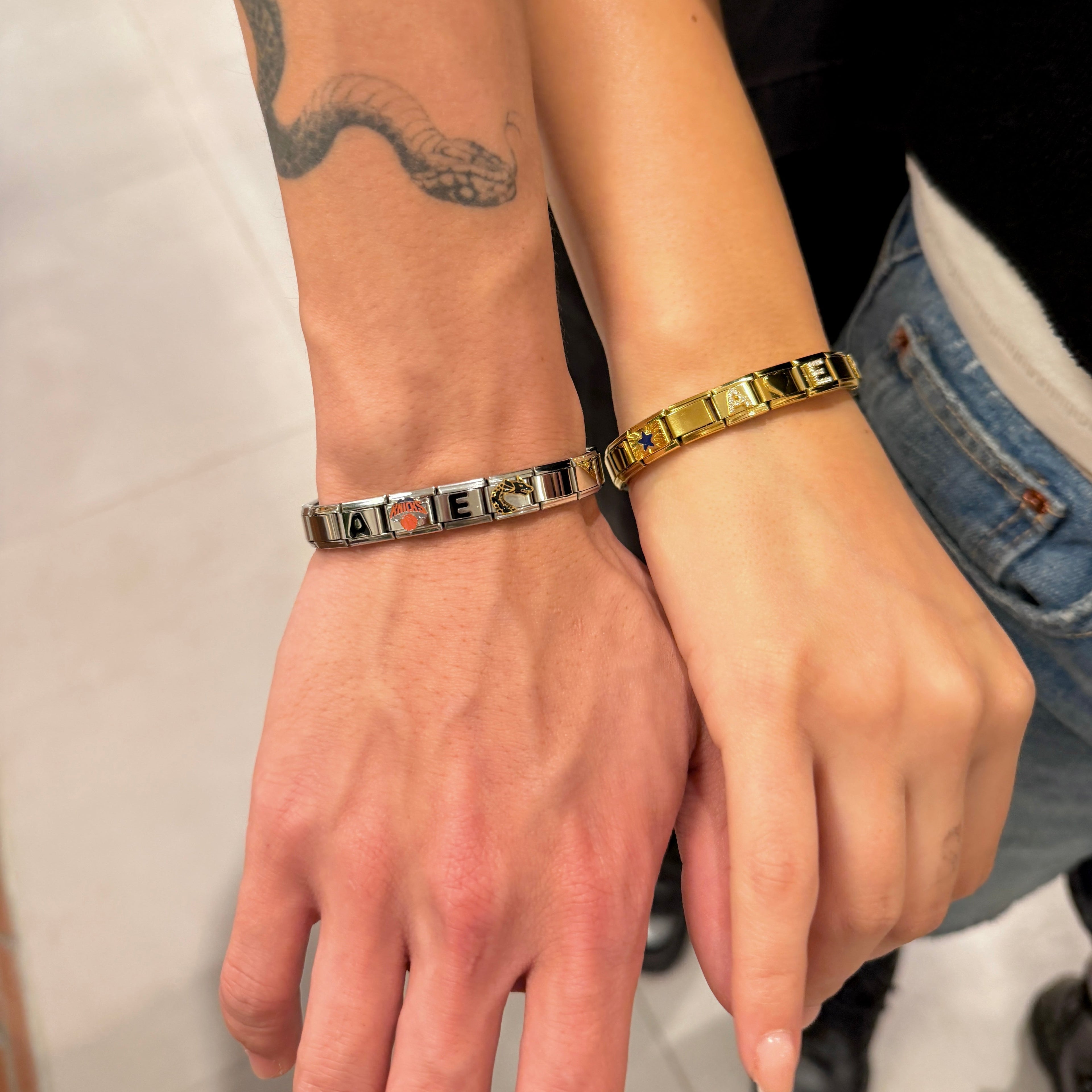 Two Italian charm bracelets worn on two people's wrist, one in silver with red and white letter accents and the other in yellow gold with white letter accents, shown on a model's wrist against a neutral indoor background.