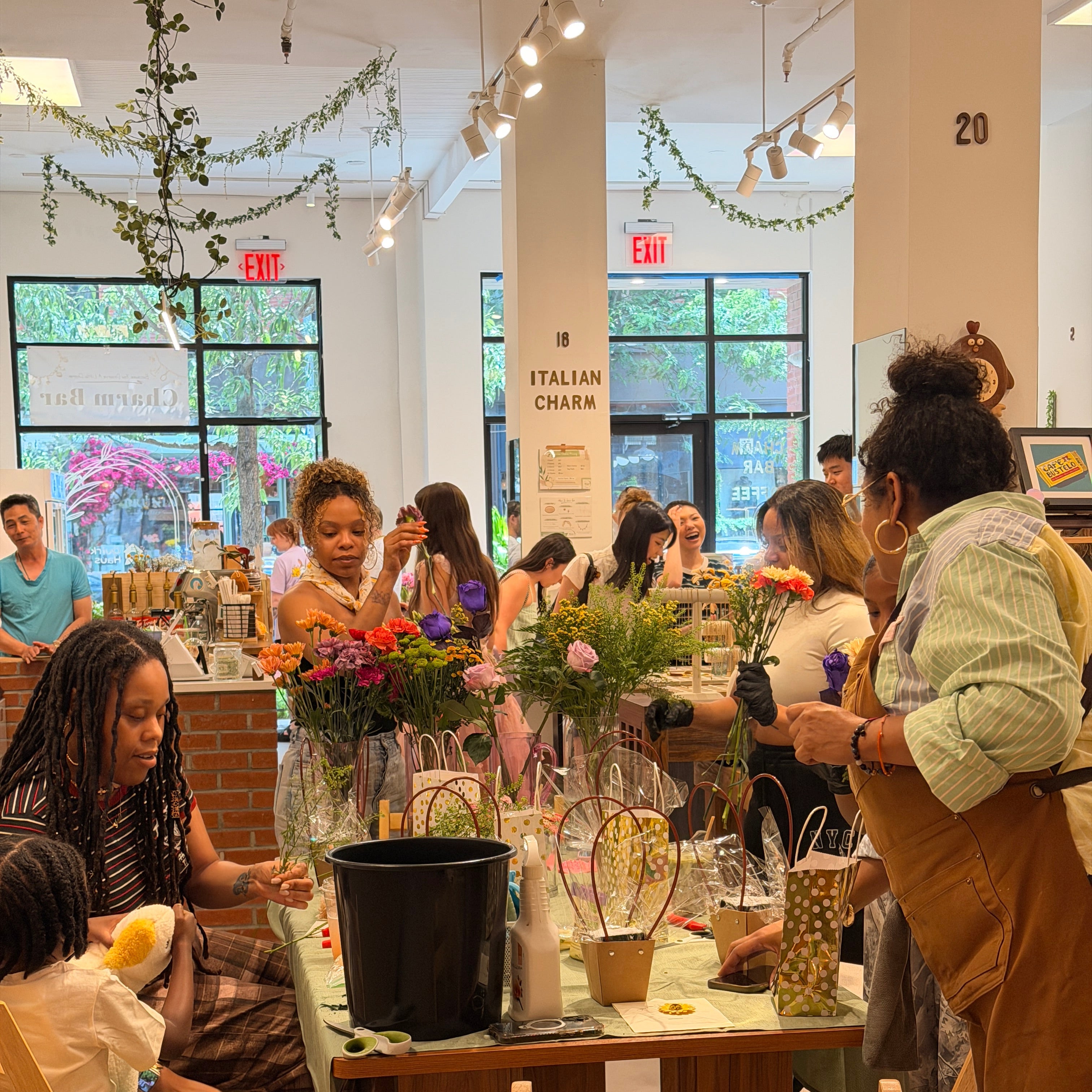 A busy indoor event or workshop scene with multiple people gathered around tables, standing, and interacting near floral arrangements and display items in a bright, modern interior with large windows.