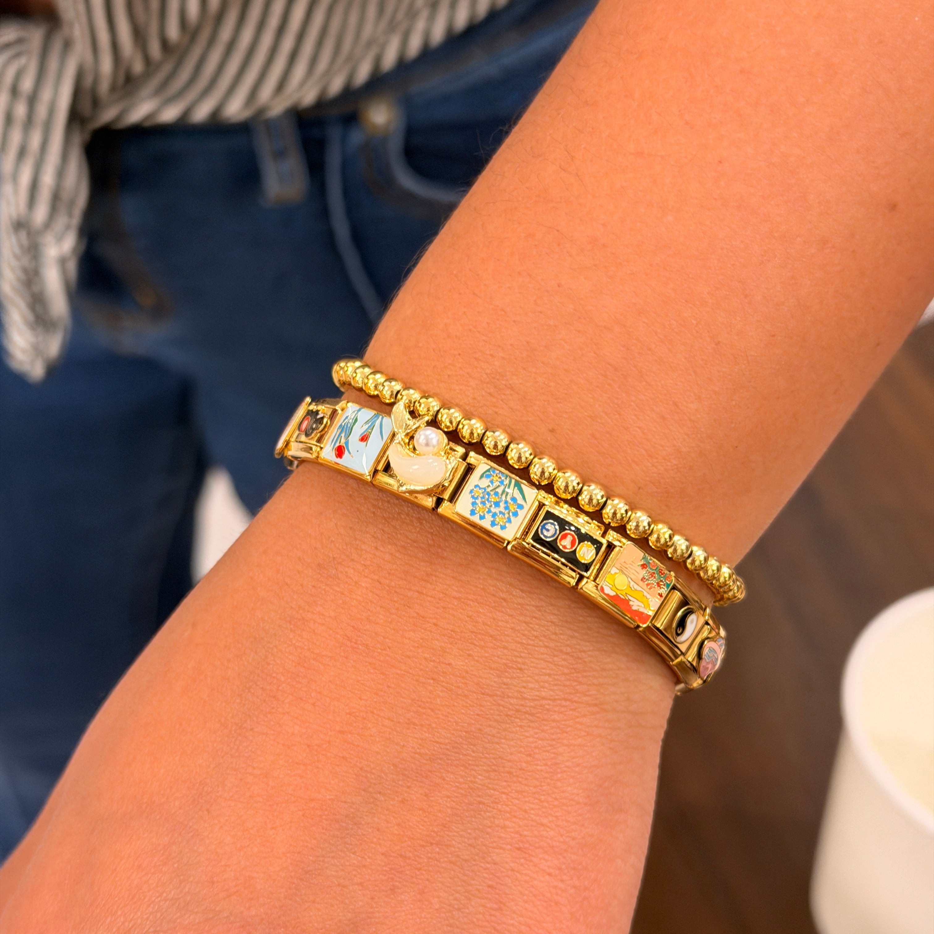 A close-up of a person's wrist wearing two gold bracelets: one beaded bracelet and one Italian charm bracelet with colorful enamel inserts featuring cartoon-style characters.