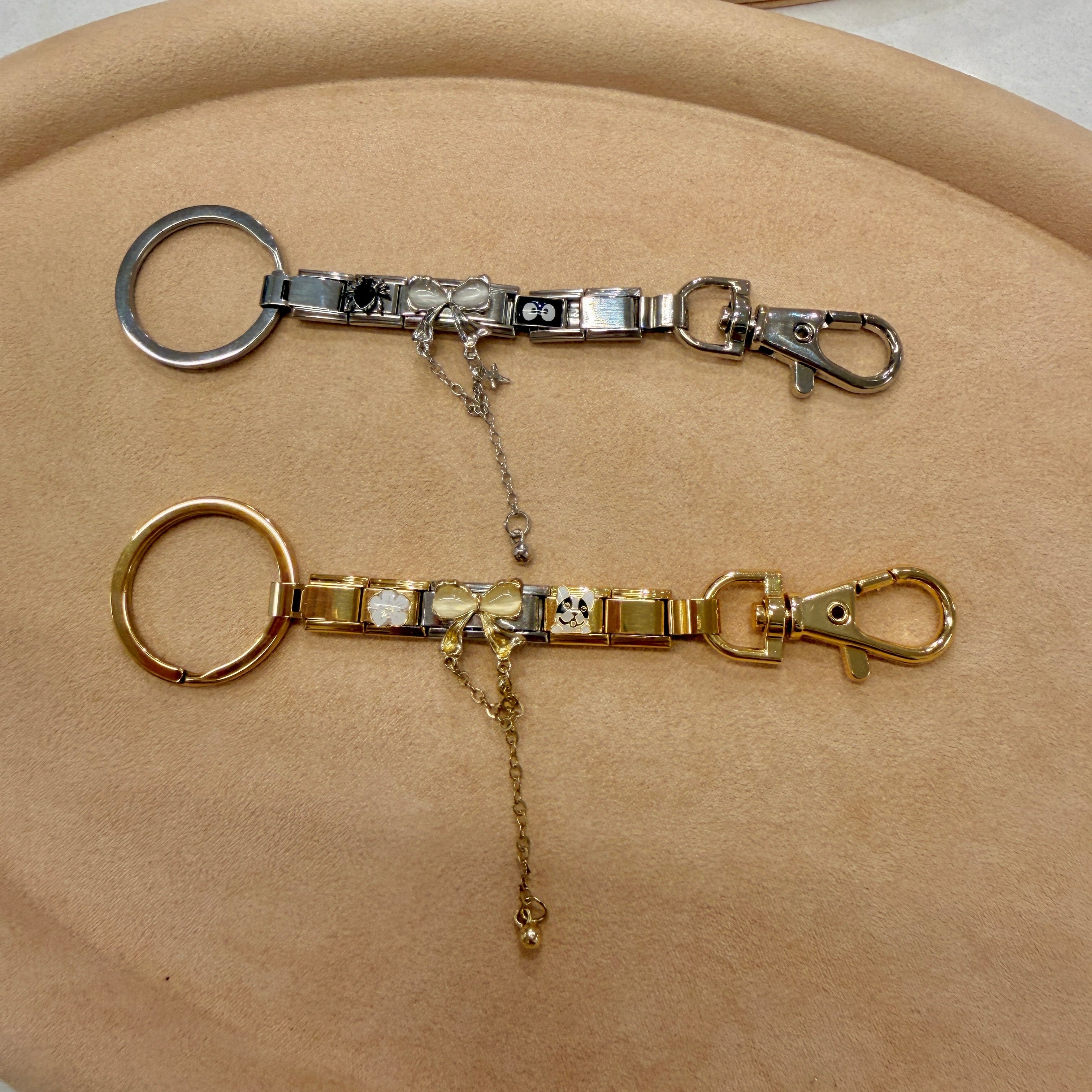 Two Italian charm keychain extensions displayed on a beige fabric surface, one in silver-tone and one in gold-tone, each with a ring, chain, and lobster clasp.