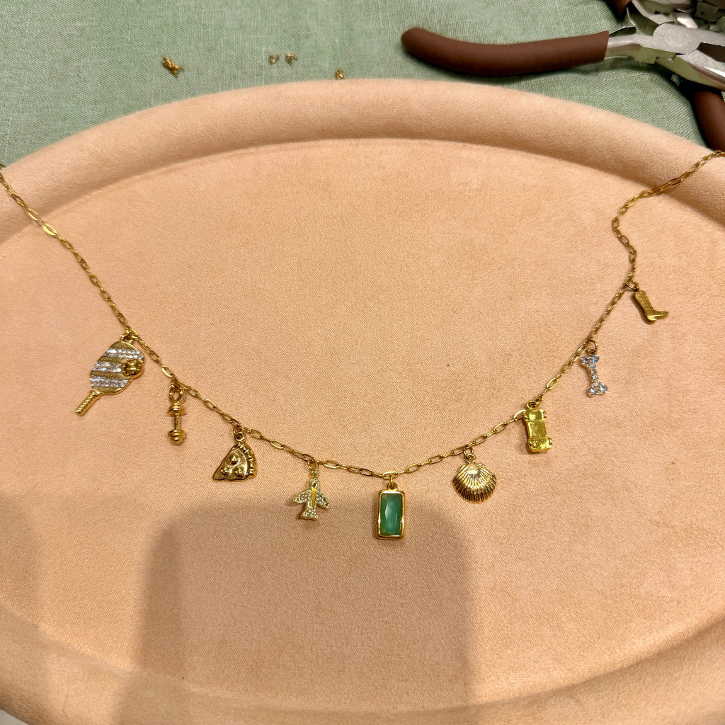 A gold charm necklace with multiple small fashion-themed charms, including a handbag, sunglasses, boots, and a green rectangular pendant, displayed on a beige jewelry stand.