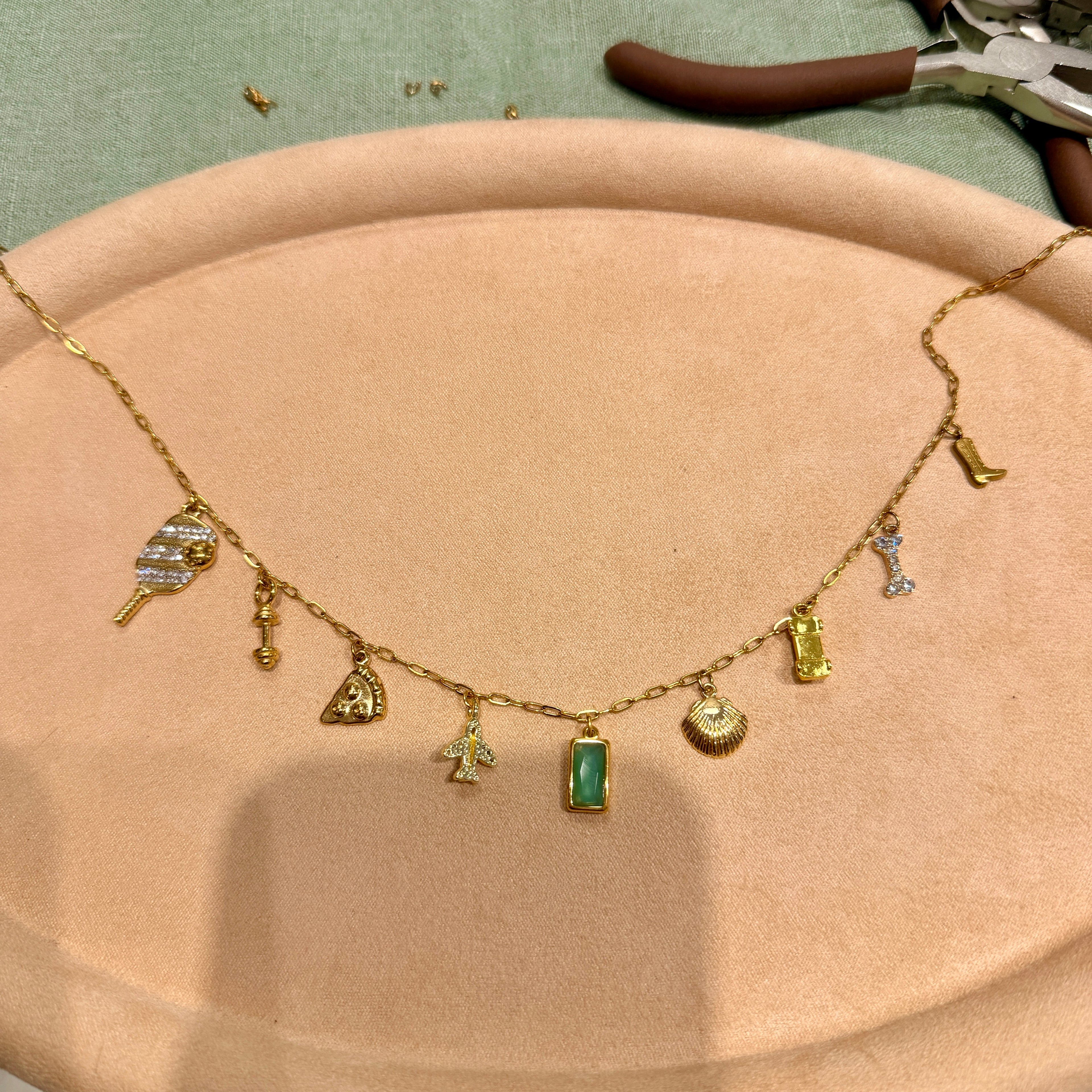 A gold charm necklace with multiple small fashion-themed charms, including a handbag, sunglasses, boots, and a green rectangular pendant, displayed on a beige jewelry stand.