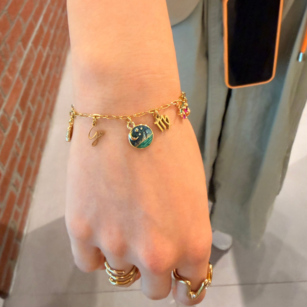 A gold charm bracelet with multiple small charms, including a green and blue marbled pendant, worn on a person's wrist. The person is also wearing gold rings, and a phone case is visible in the background.
