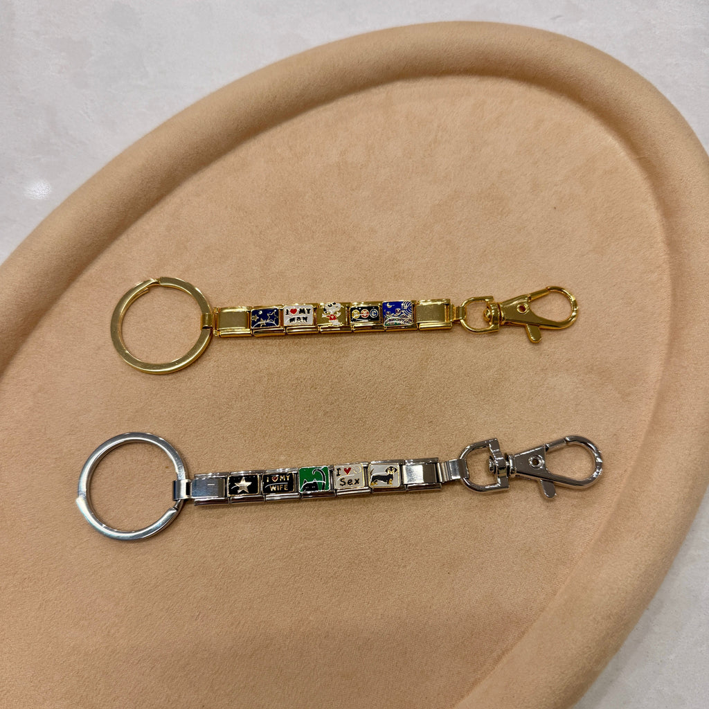 Two keychains with decorative Italian charms, one gold-tone and one silver-tone, displayed on a beige oval tray.