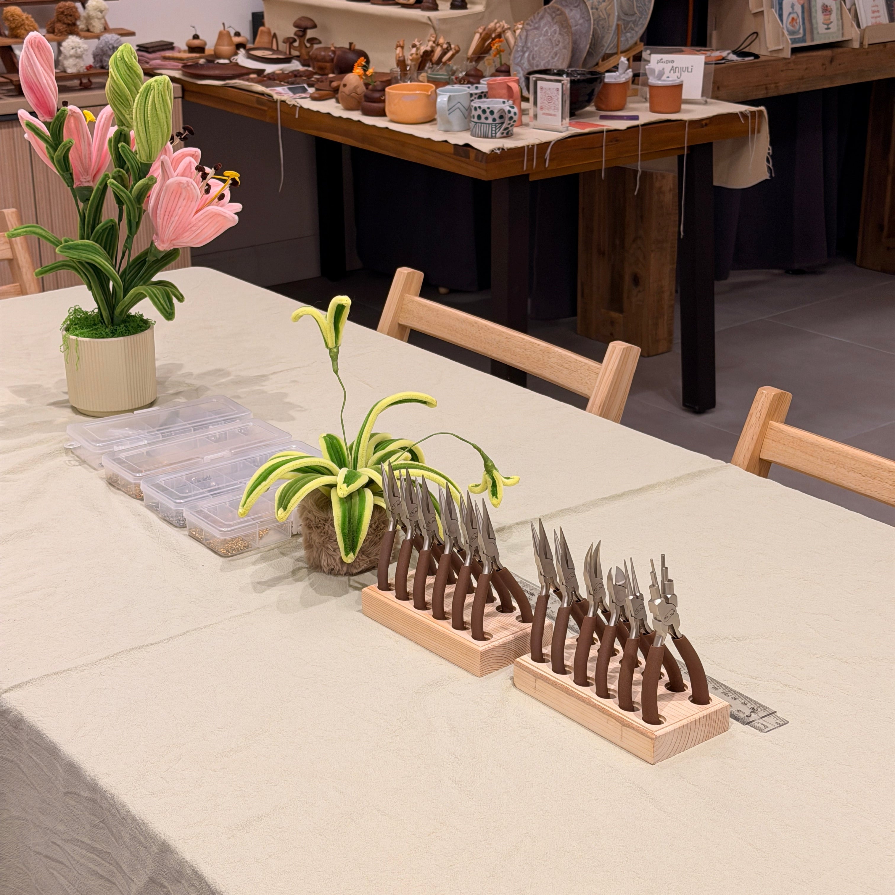 A set of kitchen knives with dark brown handles and silver blades, organized in a wooden display stand on a light-colored table. The photo is taken from an angled overhead perspective in what appears to be a rustic kitchen or shop setting, with decorative pink lilies and other items visible in the background.