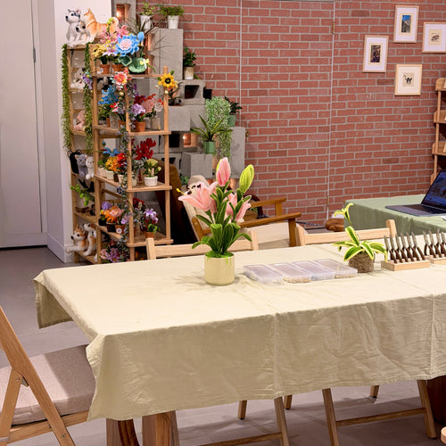 A light beige linen tablecloth draped over a wooden dining table in a cozy room with a brick wall, wooden chairs, and a vase of pink tulips in the center.