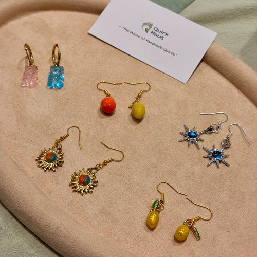 A collection of handmade earrings displayed on a beige fabric surface next to a white card from 'Quick Haus'. The earrings feature various colorful charms including pink and blue gummy bears, orange and yellow beads, gold sunflowers, and silver starbursts with blue stones.