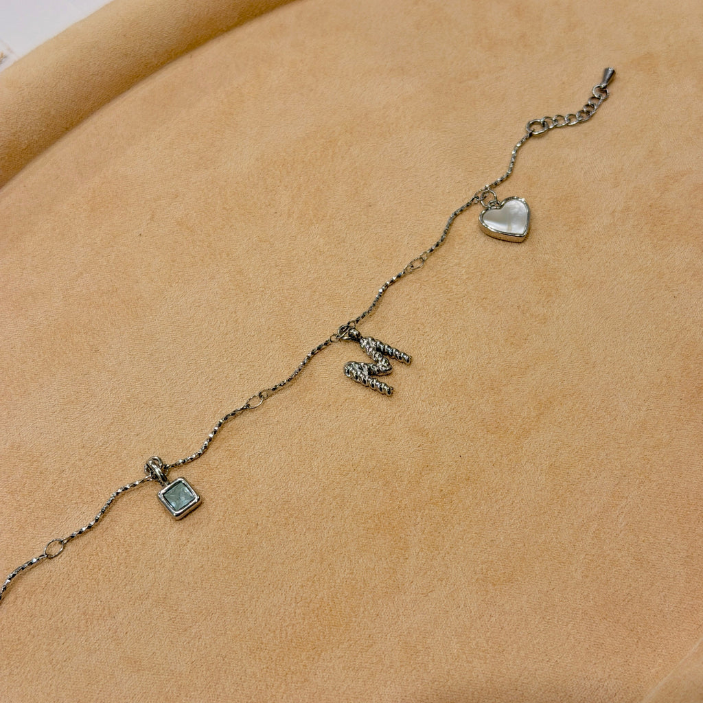 A silver-tone initial necklace spelling 'M' with a small heart charm and a square pendant, displayed on a beige velvet jewelry tray.