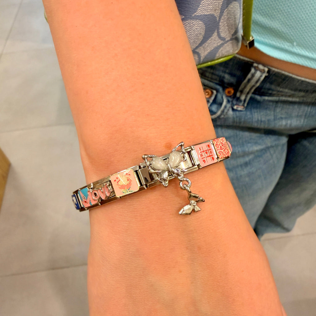 A close-up of a person's wrist wearing a silver italian charm bracelet with multiple colorful nameplates and a small dangling charm, with jeans and a patterned top visible in the background.
