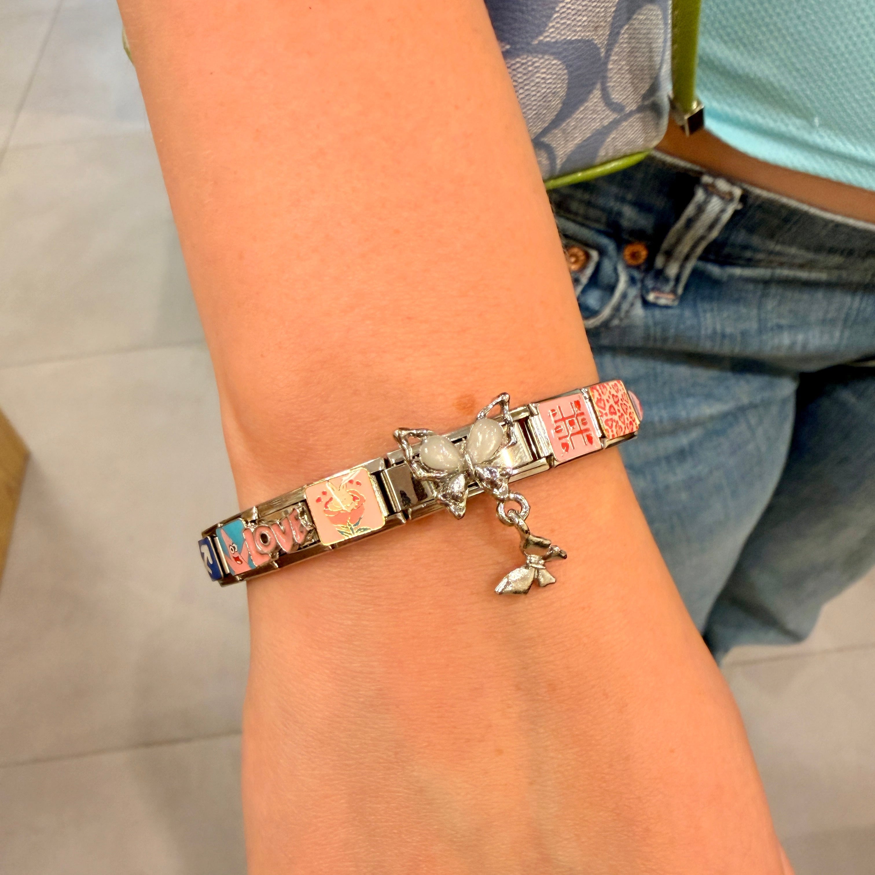 A close-up of a person's wrist wearing a silver italian charm bracelet with multiple colorful nameplates and a small dangling charm, with jeans and a patterned top visible in the background.