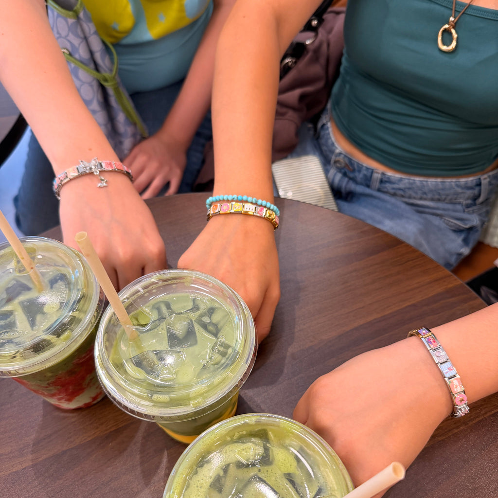 Three iced matcha green drinks in clear plastic cups with lids and straws, arranged on a dark wooden table with multiple people's hands and torsos visible around them.