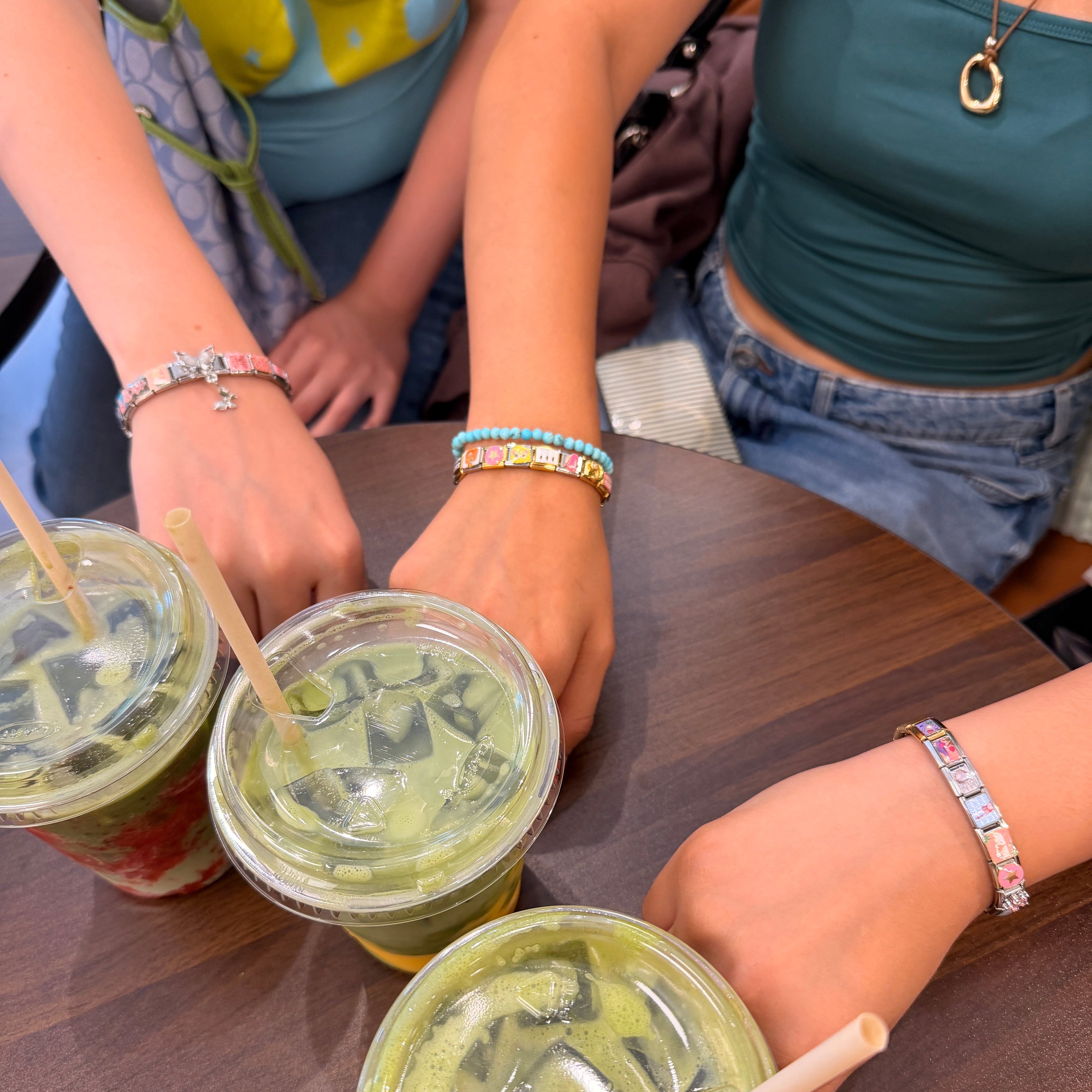 Three iced matcha green drinks in clear plastic cups with lids and straws, arranged on a dark wooden table with multiple people's hands and torsos visible around them.