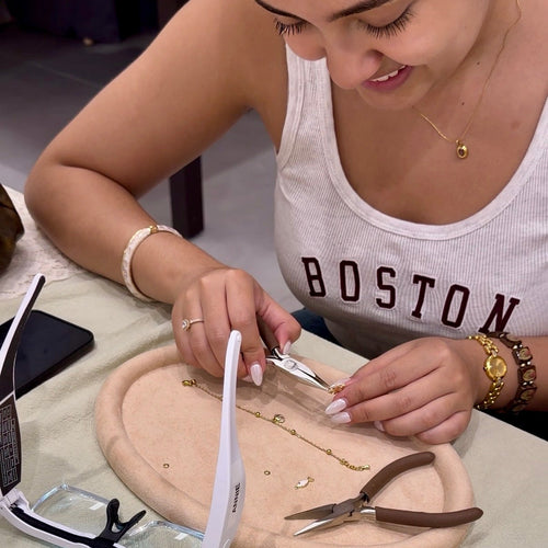 A woman is seated at a table, using tweezers to work on a delicate piece of jewelry on a small beige stand. She is wearing gold jewelry, and a pair of white-framed glasses and pliers are on the table next to her.