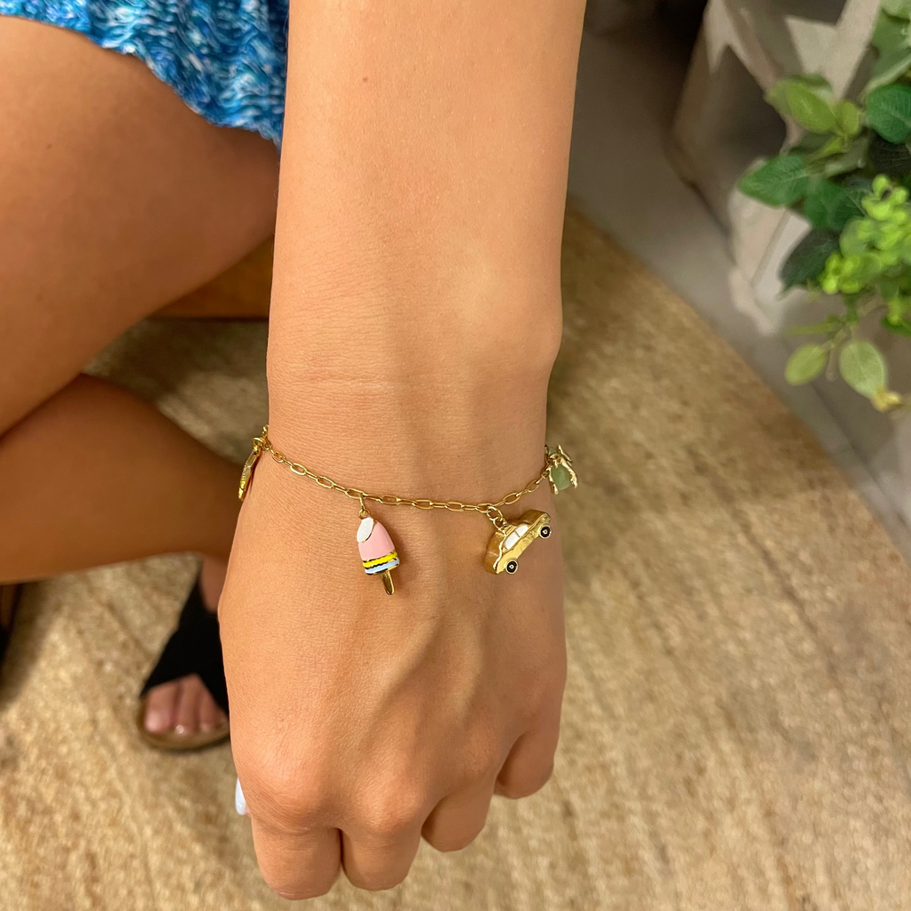 A gold-toned charm bracelet worn on a person's wrist, featuring a small pink and yellow ice cream cone charm and a small green leaf charm, shown in a casual indoor setting.